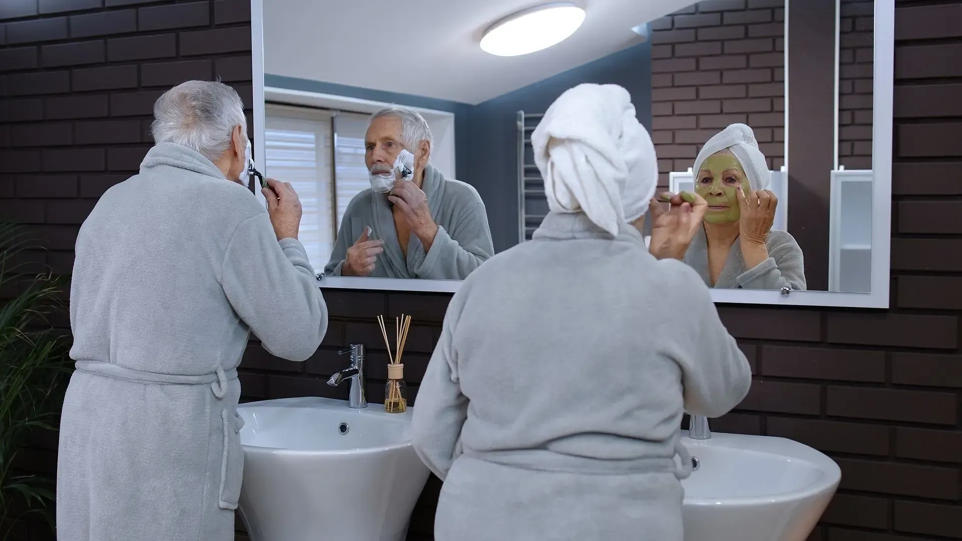 Senior man shaving at bathroom mirror while woman applies facial mask, both wearing robes and towels
