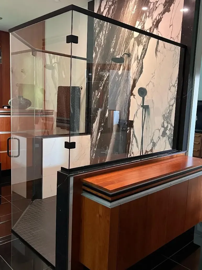 Modern bathroom display featuring frameless glass shower enclosure with marble-look tile walls, multiple showerheads, and towel bars, with wood cabinetry base in showroom setting
