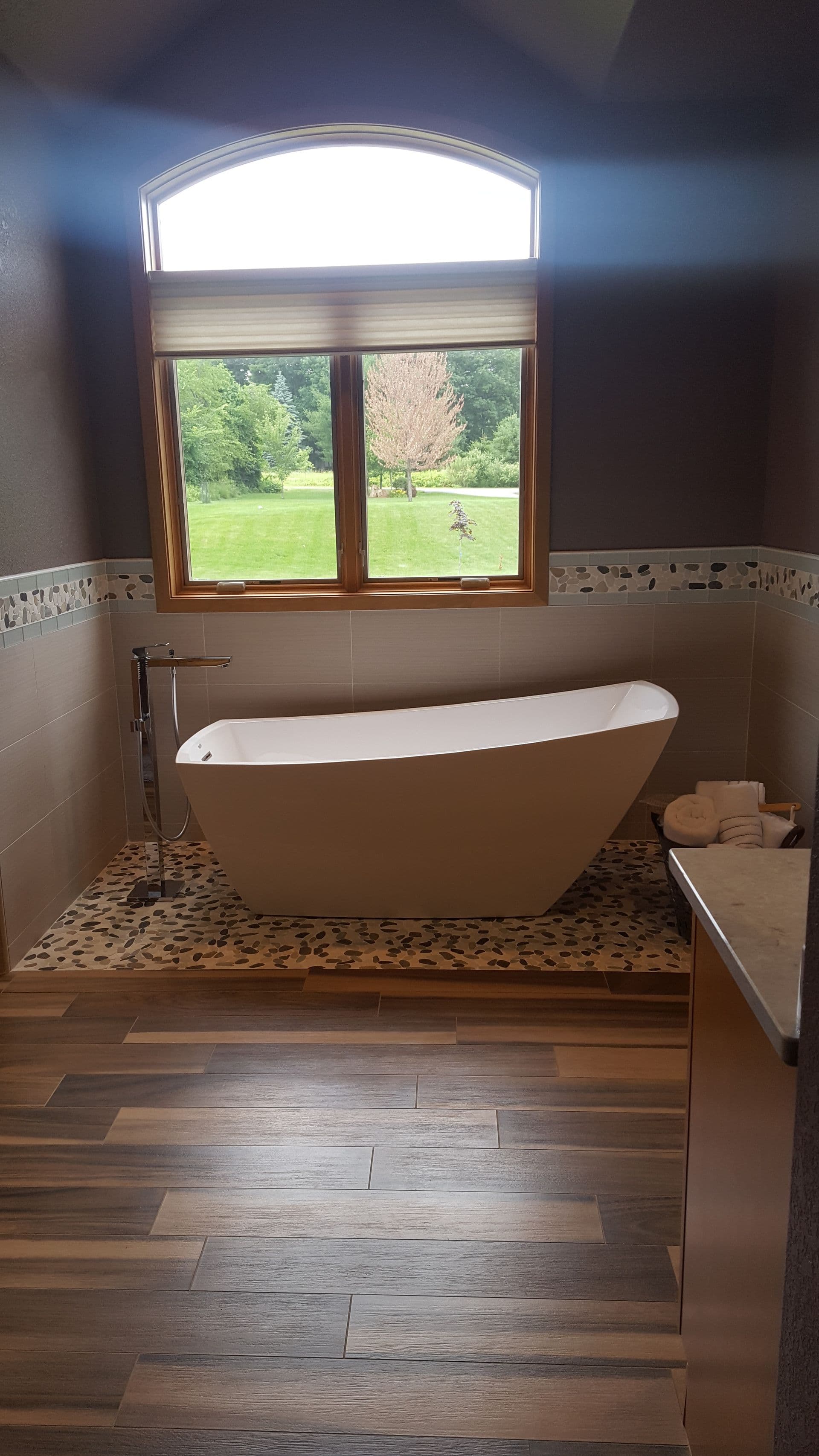 Luxury bathroom with freestanding white soaker tub on pebble tile floor, arched window overlooking green lawn and trees, wood-look plank flooring, and towel storage