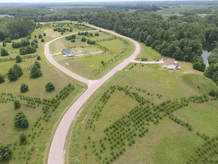 Aerial view of a rural estate with a circular driveway, central house, tree-lined grounds, and connecting access road through farmland with water visible on the right edge